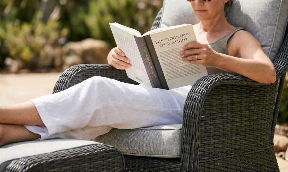 An intimate photograph of a relaxed person, fully ensconced and reading an open hardcover book titled "THE GEOGRAPHY OF SUNLIGHT" while comfortably settled within a charcoal grey all-weather resin wicker outdoor reading armchair with textured grey cushions. The view is mostly from the waist down, showing the person, who wears sunglasses and linen pants, with their legs extended and supported by a matching ottoman. Hands hold the book naturally. Focus is incredibly sharp on the tactile textures of the wicker, the fabric, the book pages, and the detailed hands holding the book. Natural sunlight illuminates the scene, highlighting the sheens and textures.