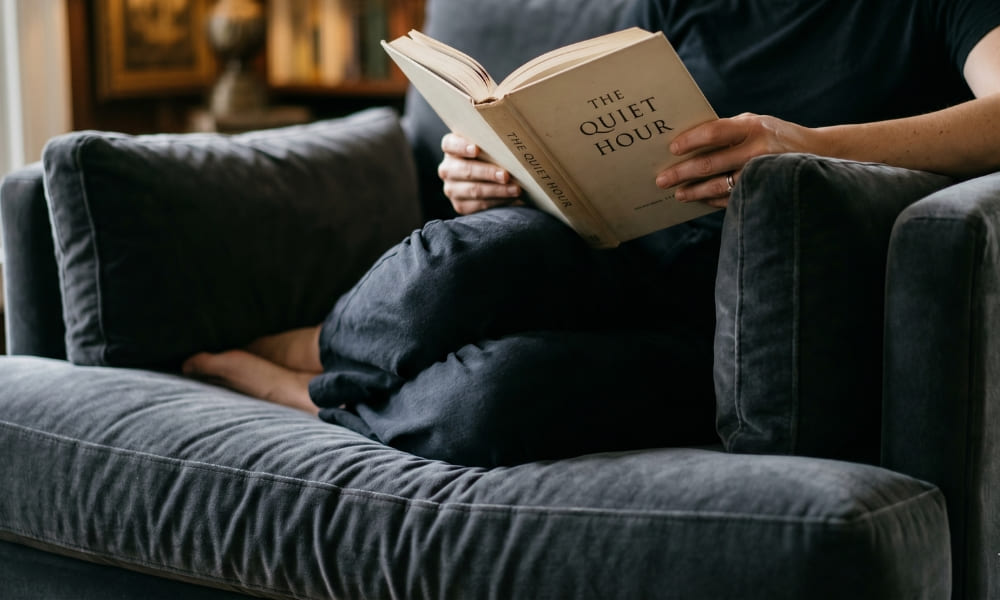 A close-up, natural-light photograph of a person seated comfortably and relaxed in a large, plush dark charcoal velvet armchair. The person, wearing dark clothing, is holding and reading an open hardcover book with the title 'THE QUIET HOUR' clearly visible on the cover and spine. The person has their legs tucked up. In the background, blurred bookshelves and soft window light create a cozy indoor reading nook atmosphere.