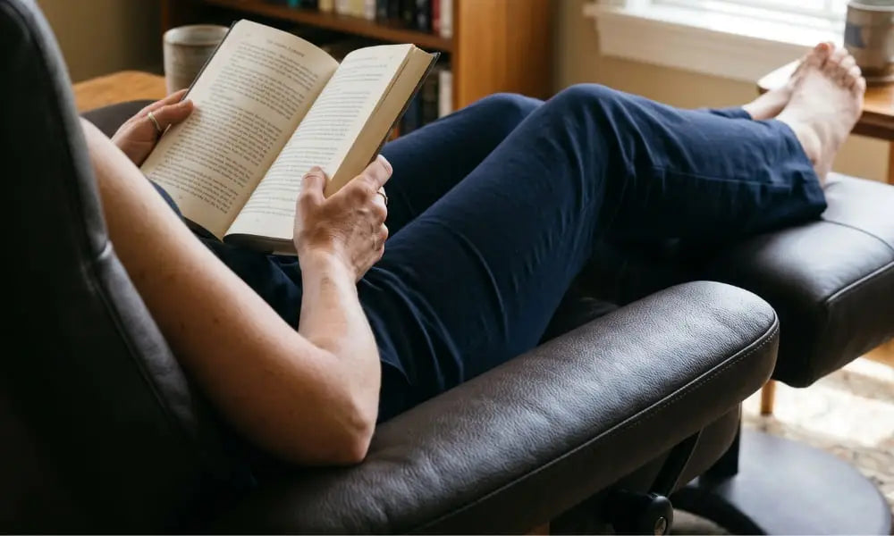 A person from the waist up is seated comfortably and relaxed in a rich, dark brown leather armchair, engrossed in reading an open hardcover book held in both hands. Their legs, wearing dark pants, are crossed and resting on a matching leather ottoman. They are barefoot. Behind them, a wooden bookshelf filled with books is visible. To the right, a wooden side table holds a ceramic mug, near natural light from a window. The floor is covered by a patterned rug.