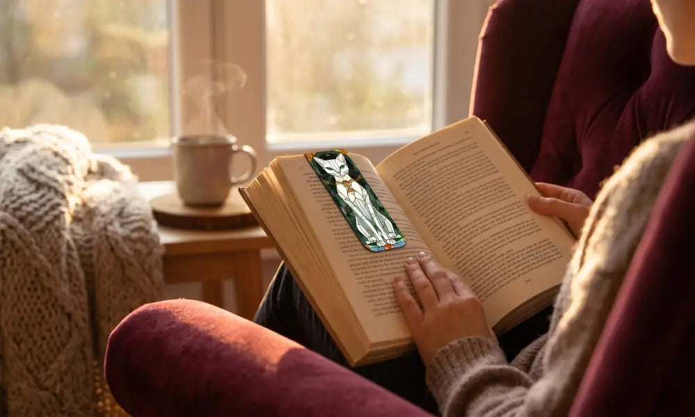 A peaceful, cozy scene of a person reading a physical book while seated in a plush burgundy velvet armchair. Sunlight from a large window during the golden hour illuminates the moment. A distinctive, stained-glass-style cat bookmark is visible. A side table with a steaming mug is in the background