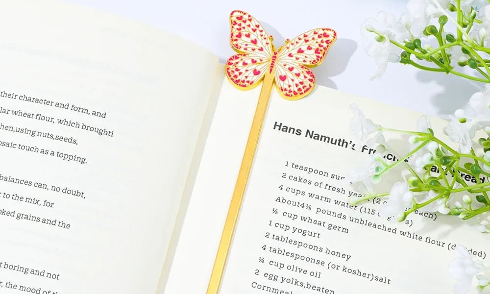 A dainty gold metal bookmark featuring a white butterfly with small pink heart patterns on its wings. The bookmark is placed inside an open cookbook recipe page, surrounded by white baby's breath flowers on a bright, clean background.