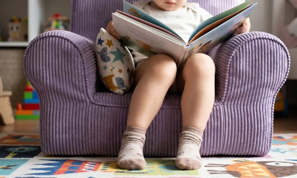 A toddler is fully ensconced and reading a large open picture book in a small, lavender corduroy kids' armchair. The child's legs and socks are visible, and a decorative patterned pillow is tucked behind them. The setting is a cozy nursery with blurred bookshelves and toys on a colorful activity rug.