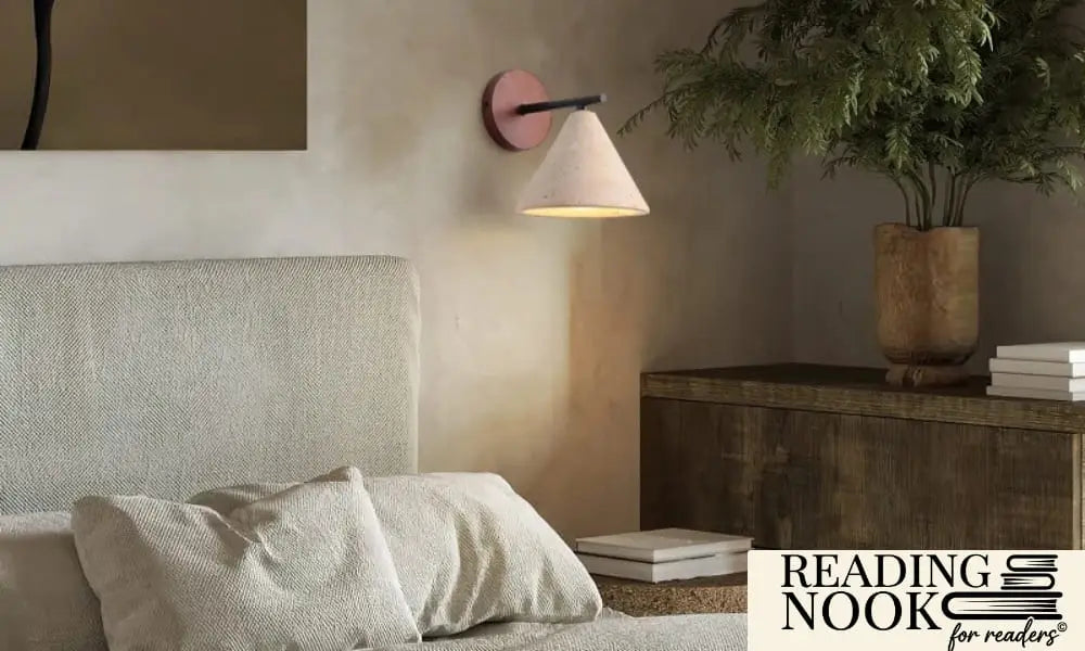 An organic modern wall sconce by Reading Nook featuring a textured stone cone shade and a round wooden wall mount. The lamp provides warm ambient lighting next to a cozy bed with beige linen headboard and pillows. A minimalist Japandi bedroom setting with earthy tones and greenery.
