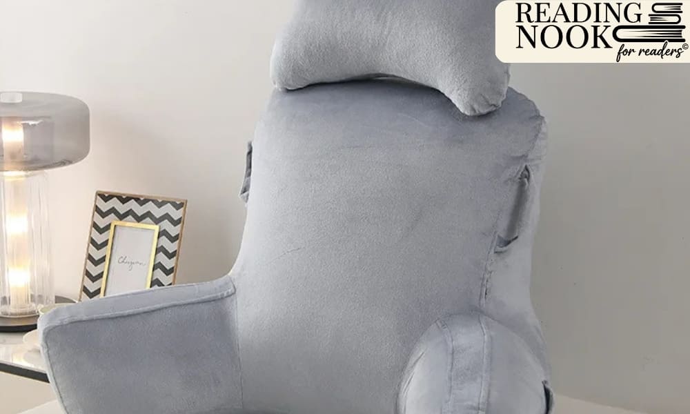 A light grey plush Reading Nook husband pillow featuring thick supportive armrests and a matching headrest. This velvety bed rest pillow is designed for back support while sitting up in bed, pictured in a modern bedroom with a lamp and decor