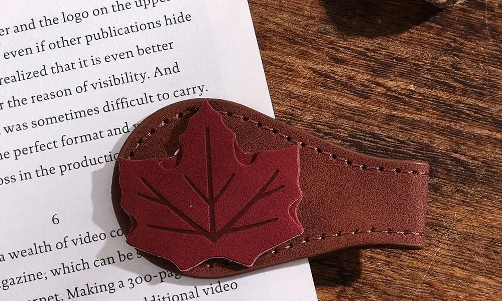 A detailed close-up photograph of a brown faux leather corner bookmark featuring a red felt maple leaf appliqué. The bookmark is clipped onto the page of an open book with visible English text, resting on a rustic wooden table next to a real acorn.