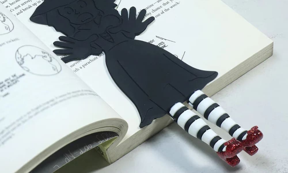 A 3D Wicked Witch bookmark shaped like a black silhouette lying inside an open book. Her legs, wearing black and white striped stockings and sparkly red ruby slippers, stick out from the bottom of the pages.